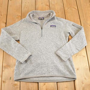 May include: A light gray fleece pullover with a quarter-zip closure and a small logo patch on the chest. The long-sleeved top has a stand-up collar and a textured appearance. The garment is laid flat on a wooden surface.