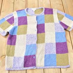 Könnte beinhalten: Eine kurzärmelige Strickjacke mit einem karierten Muster in den Farben Lila, Blau, Grün und Beige. Die Strickjacke hat einen Knopfschließe.