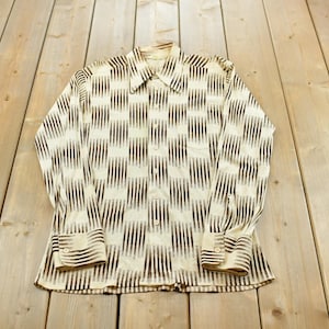 May include: A long-sleeve button-down shirt featuring a cream-colored base with a repeating vertical pattern of dark brown, blurred, brushstroke-like stripes. The shirt has a pointed collar, a front button placket, and buttoned cuffs on the sleeves.