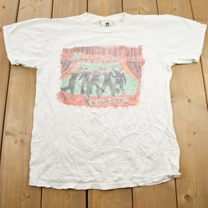 Op de afbeelding: Wit T-shirt met een vervaagde print van een band op het podium. De print toont vijf figuren in zwart-wit, met de bandnaam "The English Beat" eronder.