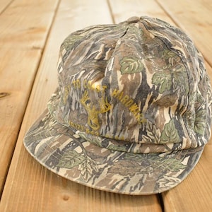 May include: Camouflage baseball cap with a curved brim. The cap features a pattern of green, brown, and tan, with a gold logo and text. The hat is likely for outdoor activities.