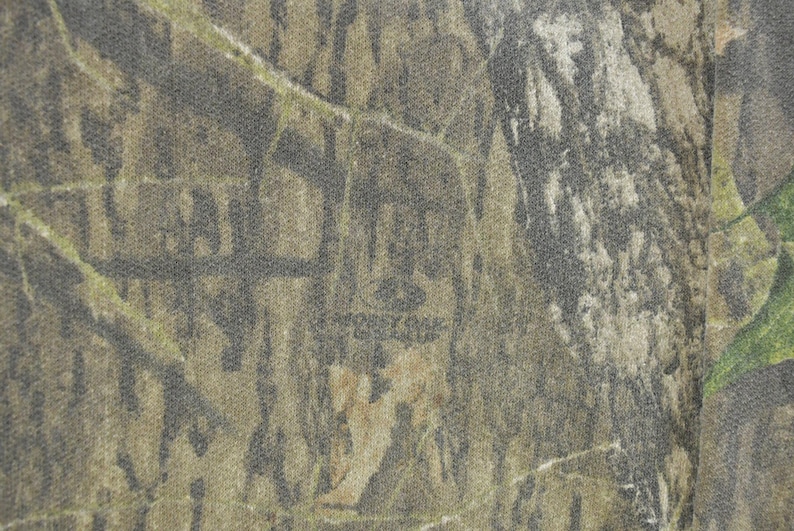 May include: Close-up of a camouflage pattern fabric. The design features a blend of brown, green, and gray hues, creating a textured, naturalistic appearance. The fabric has a subtle, mottled effect, with the "Mossy Oak" logo visible.
