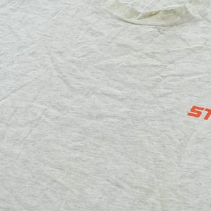 Op de afbeelding: Een lichtgrijs gem&ecirc;leerd t-shirt met een ronde hals en het oranje STIHL-logo op de rechterkant van de borst. De stof heeft een textuur met een gespikkeld uiterlijk en vertoont zichtbare kreukels over het oppervlak van het kledingstuk.