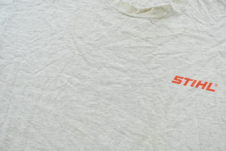 Op de afbeelding: Een lichtgrijs gem&ecirc;leerd t-shirt met een ronde hals en het oranje STIHL-logo op de rechterkant van de borst. De stof heeft een textuur met een gespikkeld uiterlijk en vertoont zichtbare kreukels over het oppervlak van het kledingstuk.