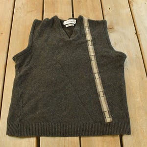 May include: Dark gray V-neck sweater vest with a decorative vertical stripe. The vest has a ribbed bottom hem and a small tag near the neckline. The stripe is a lighter shade of gray with a repeating pattern.