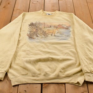 May include: A pale yellow long-sleeve sweatshirt featuring a central graphic print of a fox standing in a snowy field near a wooden wagon wheel and a red barn. The garment has a crew neckline, ribbed cuffs, and a ribbed hemline at the bottom.