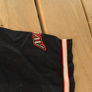 Op de afbeelding: Zwarte mesh stof met het woord "AVI" in rode en witte borduursels. Een verticale streep met rode en witte accenten loopt langs de rand van de stof. De stof maakt waarschijnlijk deel uit van een sportshirt of sportkleding.