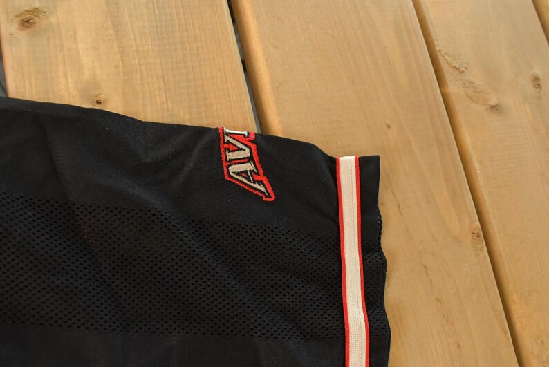 Op de afbeelding: Zwarte mesh stof met het woord "AVI" in rode en witte borduursels. Een verticale streep met rode en witte accenten loopt langs de rand van de stof. De stof maakt waarschijnlijk deel uit van een sportshirt of sportkleding.