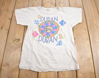May include: White t-shirt with a colorful graphic design featuring the band name "Duran Duran" and a stylized eye surrounded by a compass rose. The design includes various symbols and the numbers "24" and "12".