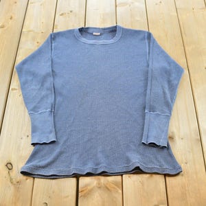 May include: A long-sleeve thermal shirt in a muted blue color. The garment features a textured waffle-knit fabric, a classic crew neckline, and wide, fitted cuffs at the end of each sleeve. The shirt has a straight hemline and a consistent blue tone.