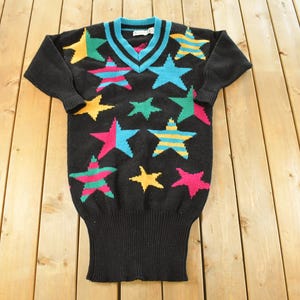 May include: Black V-neck sweater with a pattern of colorful stars. The stars are in shades of yellow, green, pink, and blue. The sweater has a ribbed hem and cuffs. The neckline is trimmed with turquoise and black.