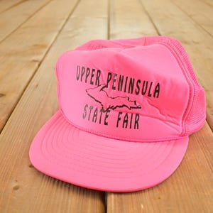 Könnte beinhalten: Leuchtend pinkfarbene Trucker-Cap mit Mesh-Rückseite und gebogenem Schirm. Auf der Vorderseite steht in Schwarz "UPPER PENINSULA STATE FAIR" mit einer Grafik der Upper Peninsula.