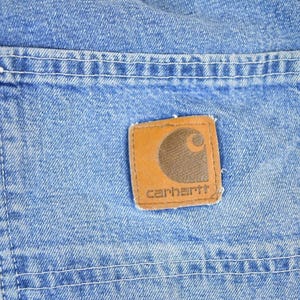 K&ouml;nnte beinhalten: Nahaufnahme von hellblauem Denim-Stoff mit einer Tasche und einem braunen Carhartt-Markenaufn&auml;her. Der Aufn&auml;her zeigt ein braunes Logo und den Markennamen in Wei&szlig;. Wei&szlig;e N&auml;hte umranden die Tasche und den Aufn&auml;her.