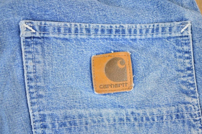 K&ouml;nnte beinhalten: Nahaufnahme von hellblauem Denim-Stoff mit einer Tasche und einem braunen Carhartt-Markenaufn&auml;her. Der Aufn&auml;her zeigt ein braunes Logo und den Markennamen in Wei&szlig;. Wei&szlig;e N&auml;hte umranden die Tasche und den Aufn&auml;her.