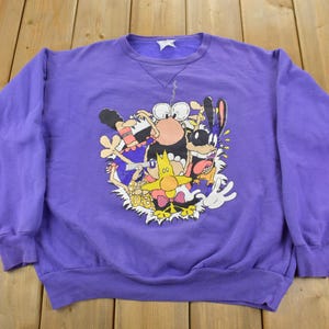 May include: Purple sweatshirt with a graphic print of cartoon characters. The characters are a yellow bird, a blue rabbit, and a black and white creature. The characters are all smiling and have large eyes.