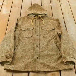 May include: A tan wool hooded jacket with two chest pockets and two lower pockets. The jacket has a button-up front, buttoned cuffs, and a drawstring hood. The jacket is laid out on a wooden surface.