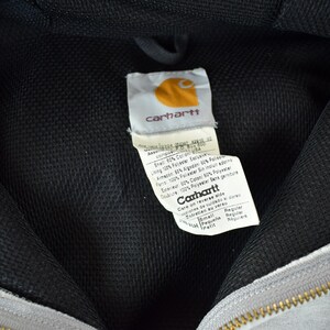 Puede incluir: Primer plano del interior de una sudadera con capucha Carhartt gris. El forro de malla negra es visible, junto con la etiqueta Carhartt. La etiqueta presenta el logotipo de Carhartt y las instrucciones de cuidado. Una cremallera dorada est&aacute; en la parte inferior.