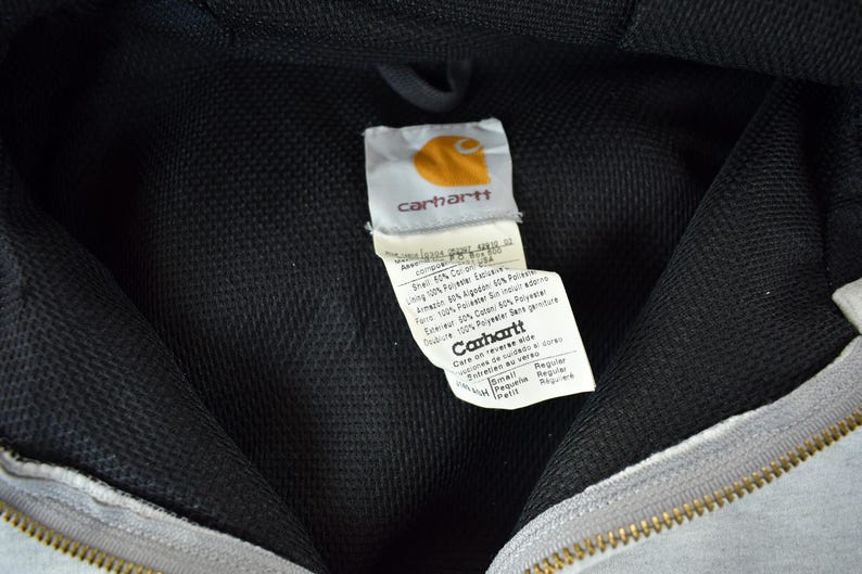 Puede incluir: Primer plano del interior de una sudadera con capucha Carhartt gris. El forro de malla negra es visible, junto con la etiqueta Carhartt. La etiqueta presenta el logotipo de Carhartt y las instrucciones de cuidado. Una cremallera dorada est&aacute; en la parte inferior.