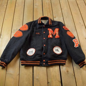 Puede incluir: Una chaqueta de béisbol negra con rayas naranjas y blancas en las mangas y el cuello. La chaqueta tiene varios parches, incluyendo una gran "M" en el pecho y un parche más pequeño en la manga izquierda que dice "Class of '54".