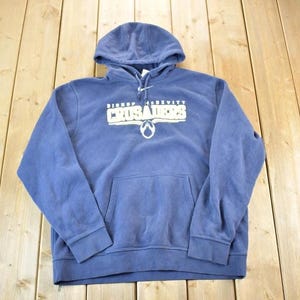 Pode incluir: Camisola azul com capuz com a inscrição branca "BISHOP McDEVITT CRUSADERS" no peito. A camisola tem um bolso frontal e um capuz com cordão. A peça de vestuário é feita de um material macio e confortável.