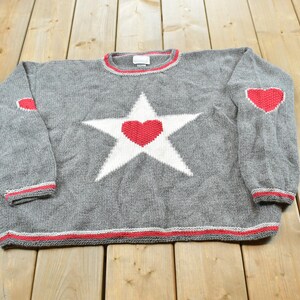 May include: A gray long-sleeve knit sweater featuring a large white star on the chest with a red heart in the center. The sweater includes red heart patches on the elbows and red and white striped trim along the crew neckline, cuffs, and hem.