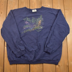 Puede incluir: Una sudadera de manga larga azul marino con un estampado gr&aacute;fico de una escena de bosque. El estampado presenta &aacute;rboles, follaje y un ciervo.