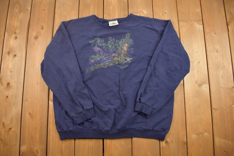 Puede incluir: Una sudadera de manga larga azul marino con un estampado gr&aacute;fico de una escena de bosque. El estampado presenta &aacute;rboles, follaje y un ciervo.
