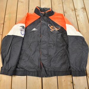 Puede incluir: Chaqueta vintage negra, naranja y blanca con cremallera completa. La chaqueta presenta un logotipo de pájaro en el pecho derecho y un parche en la manga izquierda. Cuello alzado, puños y cintura elásticos.