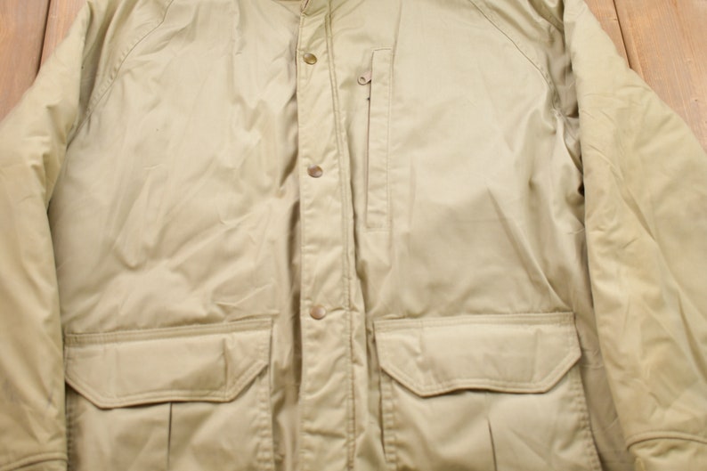 May include: A beige jacket with a button-up front, two large flap pockets, and a zippered pocket on the left side.