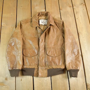 May include: A vintage brown leather jacket with a collared neckline, two front pockets, and ribbed cuffs. The jacket has a weathered appearance, suggesting age and wear. The jacket has a label that reads "Air Force".