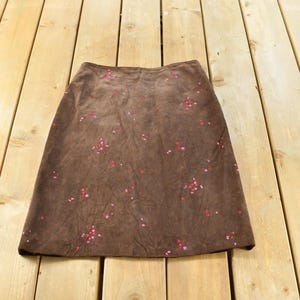 May include: A knee-length brown skirt featuring a scattered pattern of small, vibrant pink floral accents. The garment has a straight-cut silhouette with a simple waistband, showcasing a soft, textured surface throughout the entire piece.