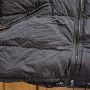 May include: Black puffer jacket with a zip fastening and a red tag on the left side. The jacket has a quilted design and a drawstring at the bottom.