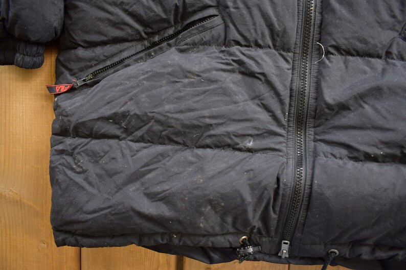 May include: Black puffer jacket with a zip fastening and a red tag on the left side. The jacket has a quilted design and a drawstring at the bottom.