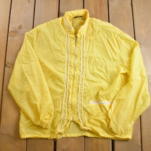 Puede incluir: Chaqueta amarilla brillante con cremallera blanca y ribete blanco en la parte delantera. La chaqueta tiene un cordón en la parte inferior y un pequeño bolsillo en el pecho. La chaqueta es de material ligero.