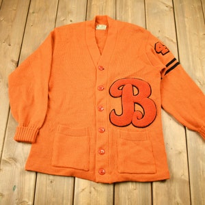 Könnte beinhalten: Orangefarbener Strickcardigan mit einem großen roten Filzbuchstaben "B" auf der Vorderseite und schwarzen und weißen Streifen an den Ärmeln. Der Cardigan hat einen V-Ausschnitt und fünf rote Knöpfe.