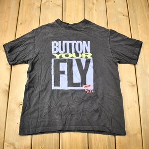 Puede incluir: Una camiseta negra con el texto "Button Your Fly" en una fuente de bloque colorida. El texto "Levi's 501" también es visible en la esquina inferior derecha.