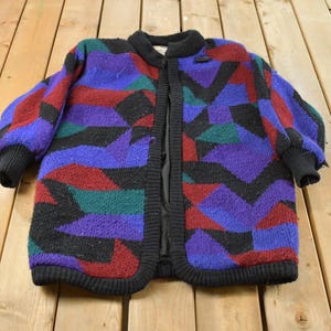 Puede incluir: Un cárdigan de punto de manga larga con un llamativo patrón geométrico en tonos morado, rojo oscuro, negro y verde azulado. La prenda tiene un cuello, puños y tapeta frontal de canalé negro, con un forro interior negro visible en la abertura.