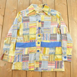 May include: A long-sleeve button-down shirt featuring a multi-colored patchwork pattern of yellow, blue, red, and white stripes and solid blocks. The shirt includes a solid blue horizontal band across the waist and five blue buttons down the front.