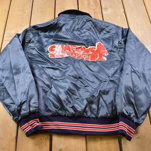 Könnte beinhalten: Vintage blaue Satinjacke mit dem Schriftzug "Shadows Saloon" in roter Schrift. Die Jacke hat einen marineblauen Kragen und Bündchen mit rot-weißen Streifen. Wahrscheinlich aus den 1980er oder 1990er Jahren.