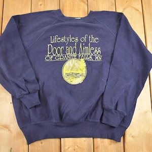 Puede incluir: Sudadera azul marino con el texto amarillo "Lifestyles of the Poor and Aimless of Granite Falls, MN". La sudadera tiene cuello redondo y mangas largas. Un gráfico circular está debajo del texto.