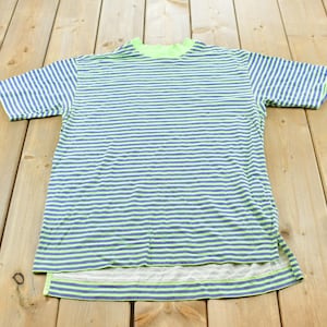 Puede incluir: Una camiseta de manga corta con un patrón de rayas horizontales en tonos azul y verde neón. La prenda tiene un cuello alto, un dobladillo recto y pequeñas aberturas laterales en la parte inferior, mostrando un diseño de rayas consistente.