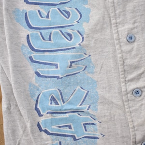 May include: A gray baseball jersey with a blue and white graphic that says "SAFERS" on the left side. The jersey has a button-up front.