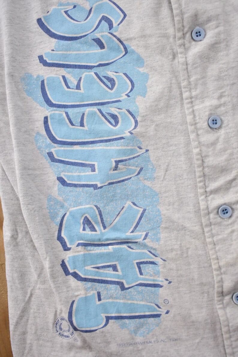 May include: A gray baseball jersey with a blue and white graphic that says "SAFERS" on the left side. The jersey has a button-up front.