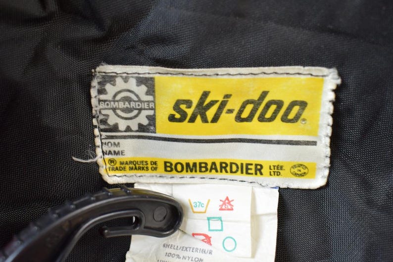 May include: A yellow and white label with the text "ski-doo" and "BOMBARDIER LTEE LTD" in black. The label also features a gear with the word "BOMBARDIER" inside.