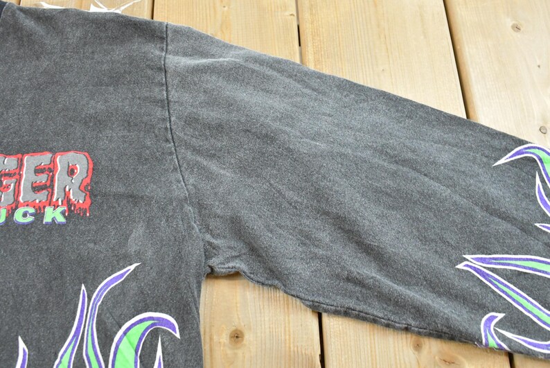 May include: Dark gray long-sleeve shirt with the word "ANGER" in red and gray, and "UCK" in green. The sleeves have a flame design in purple and green. The shirt is made of a soft, comfortable material.