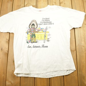 May include: White t-shirt with a cartoon illustration of a man wearing a sombrero and holding a drink. The text on the shirt reads "I'm Quiet I'm Happy I'm Handsome I don't give a Chit!!" and "San Antonio, Texas".