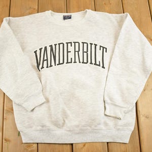 May include: A light gray crewneck sweatshirt with the word "VANDERBILT" arched across the chest in black lettering. The sweatshirt is made of a soft material and has long sleeves and ribbed cuffs and hem. The garment is laid flat on a wooden surface.