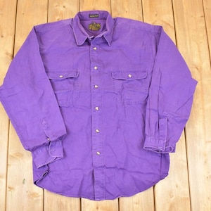 May include: A long-sleeved, button-down shirt in a vibrant purple hue. The shirt features two chest pockets, buttoned cuffs, and a classic collar. The garment is laid flat on a wooden surface.