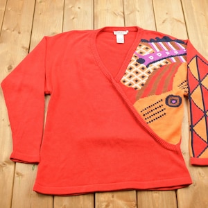 May include: A red long-sleeved sweater with a geometric pattern on the right sleeve and a colorful abstract design on the right side of the front. The sweater has a V-neck.