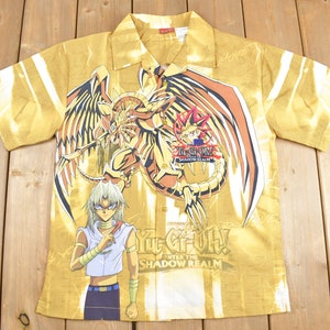May include: A gold short-sleeved button-up shirt with a graphic print of a dragon and characters from the Yu-Gi-Oh! anime series. The shirt has the text "Yu-Gi-Oh! Enter the Shadow Realm" printed on the front.
